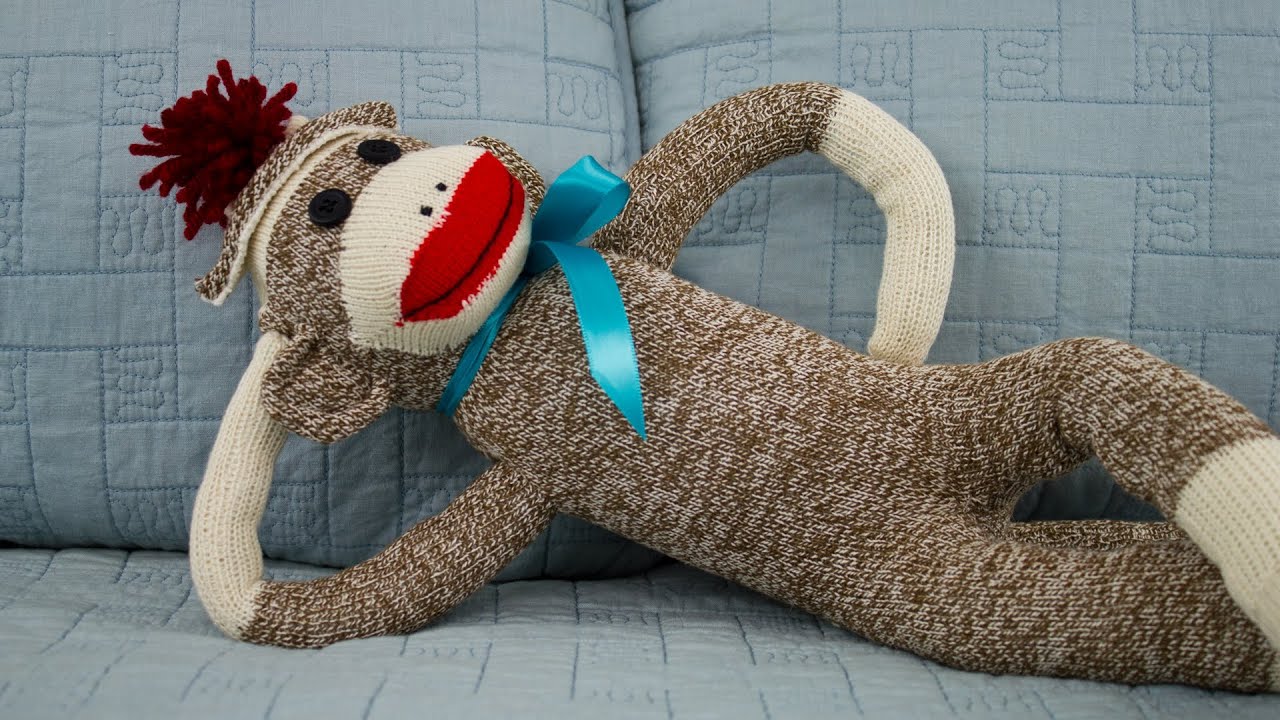 A classic handcrafted sock monkey, wearing a bright blue satin bow tie and red tassel cap, is lounging casually on a blue quilted sofa and looking back at the camera.